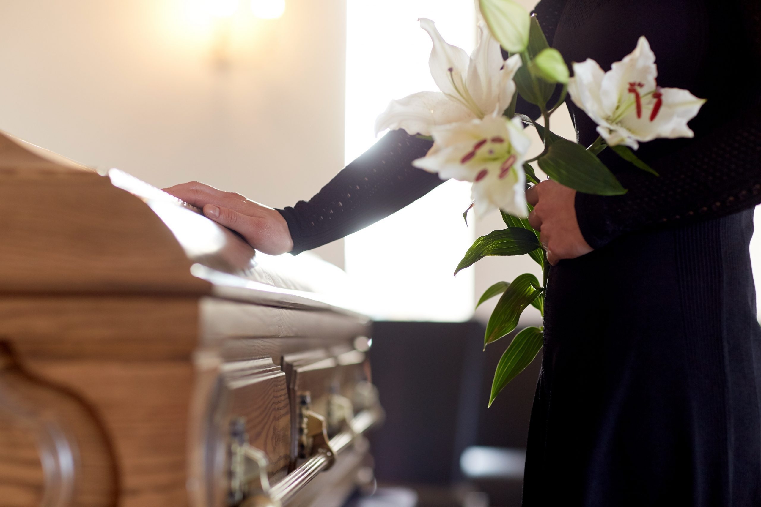 Two people standing at a wooden coffin, one resting their hand on top of it and the other holding a lilies