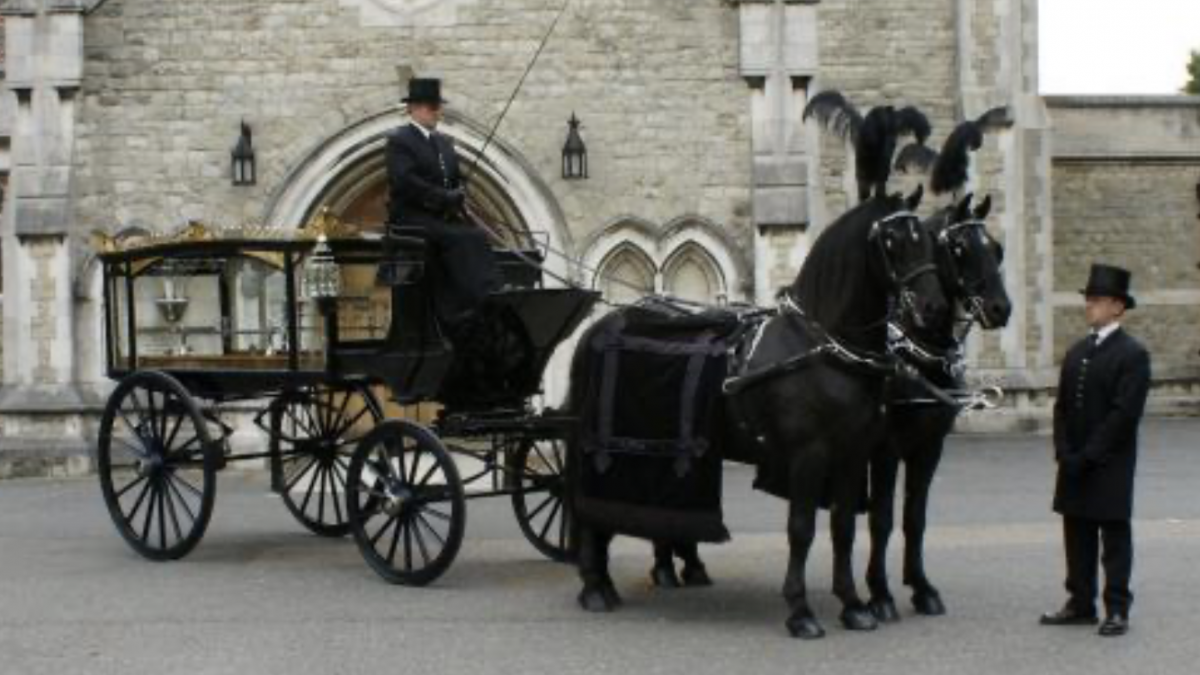 Traditional horse drawn funeral carriage. Two black horses with decorated in large black feathers and two undertakers dressed in black with top hats.